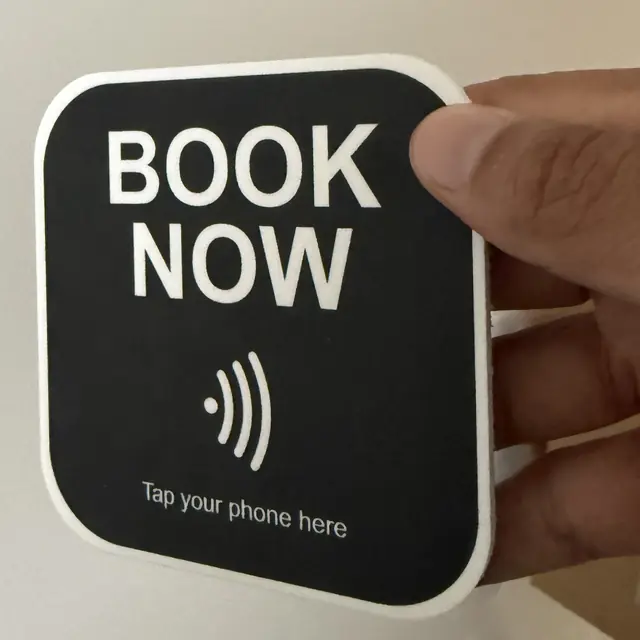 Joe's hand holding a real KEY BOOK NOW plaque. It says: Tap your phone here.