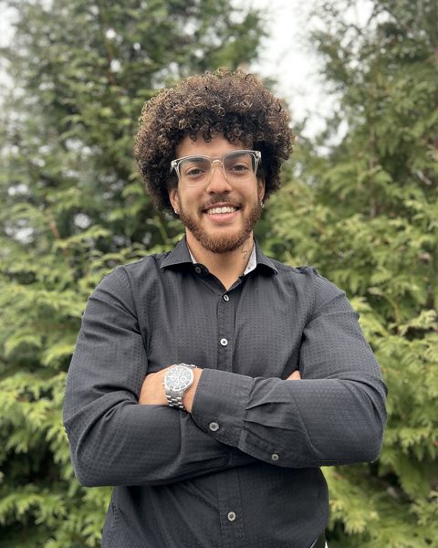 Joe, the founder of Get Key Solutions, smiling with his arms crossed in front of evergreen trees.